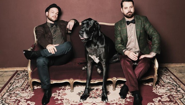 Zwei Männer und eine schwarze Dogge sitzen auf einem alten Sofa